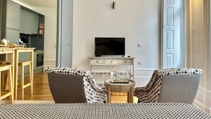 A living room with a TV on a wooden cabinet, two patterned armchairs, a small round table with a bottle, and a kitchen in the background.
