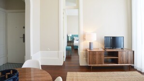 A hotel room with a wooden TV stand, a flat-screen TV, a lamp, a small table, and a chair. There is a door leading to another room and a staircase in the background. - Exceptional Two-Bedroom, Apart in Bolhão Market (Oporto)