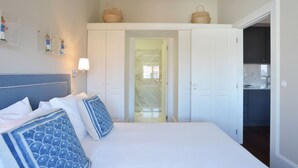 A bedroom with a bed, white walls, a bathroom door, and a kitchen area visible through an open door. - Two-Bedroom Family Ap in Bolhão Market (Oporto)