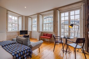 A hotel room with a bed, a sofa, a red chair, a table with a lamp, and large windows offering a city view. - Superb Studio with Balcony at the heart of Porto (Oporto)