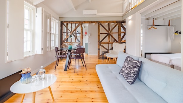 A modern living room with a wooden floor, a light blue sofa, a dining area with a table and chairs, and a staircase with wooden railings. - Amazing Duplex Two-Bedroom Apart with Views (Oporto)