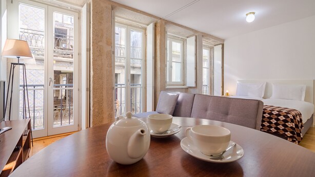 A hotel room with a wooden table set with a teapot and cups, a sofa, and a bed with a checkered blanket.