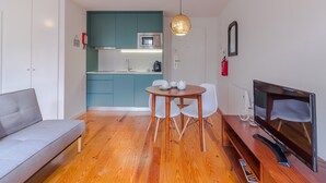 Un salon moderne avec un canapé gris, un plancher en bois, une table à manger avec des chaises, un téléviseur et un coin cuisine doté de meubles turquoise et d’un four à micro-ondes.