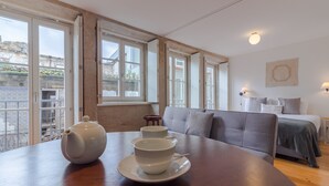 Une pièce avec une table en bois, un canapé et un lit. Il y a des fenêtres d’où l’on peut voir les bâtiments à l’extérieur.