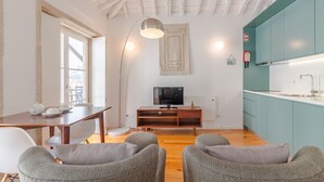 Ein modernes Wohnzimmer mit Holzboden, einem Esstisch mit Stühlen, einem Fernseher auf einem Holzbodenständchen und einer Küche mit türkisfarbenen Schränken.
