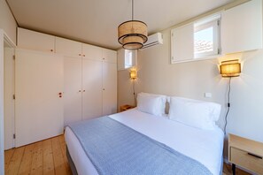 Un dormitorio con cama, armario, mesita de noche, lámpara de pared y ventana con persianas.