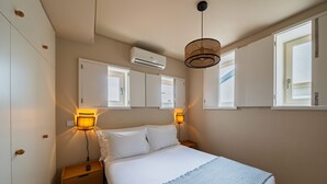 A bedroom with a bed, bedside lamps, an air conditioner, and a window with blinds.