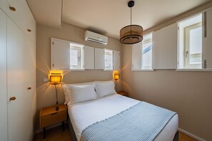 A bedroom with a bed, bedside lamps, an air conditioner, and a window.