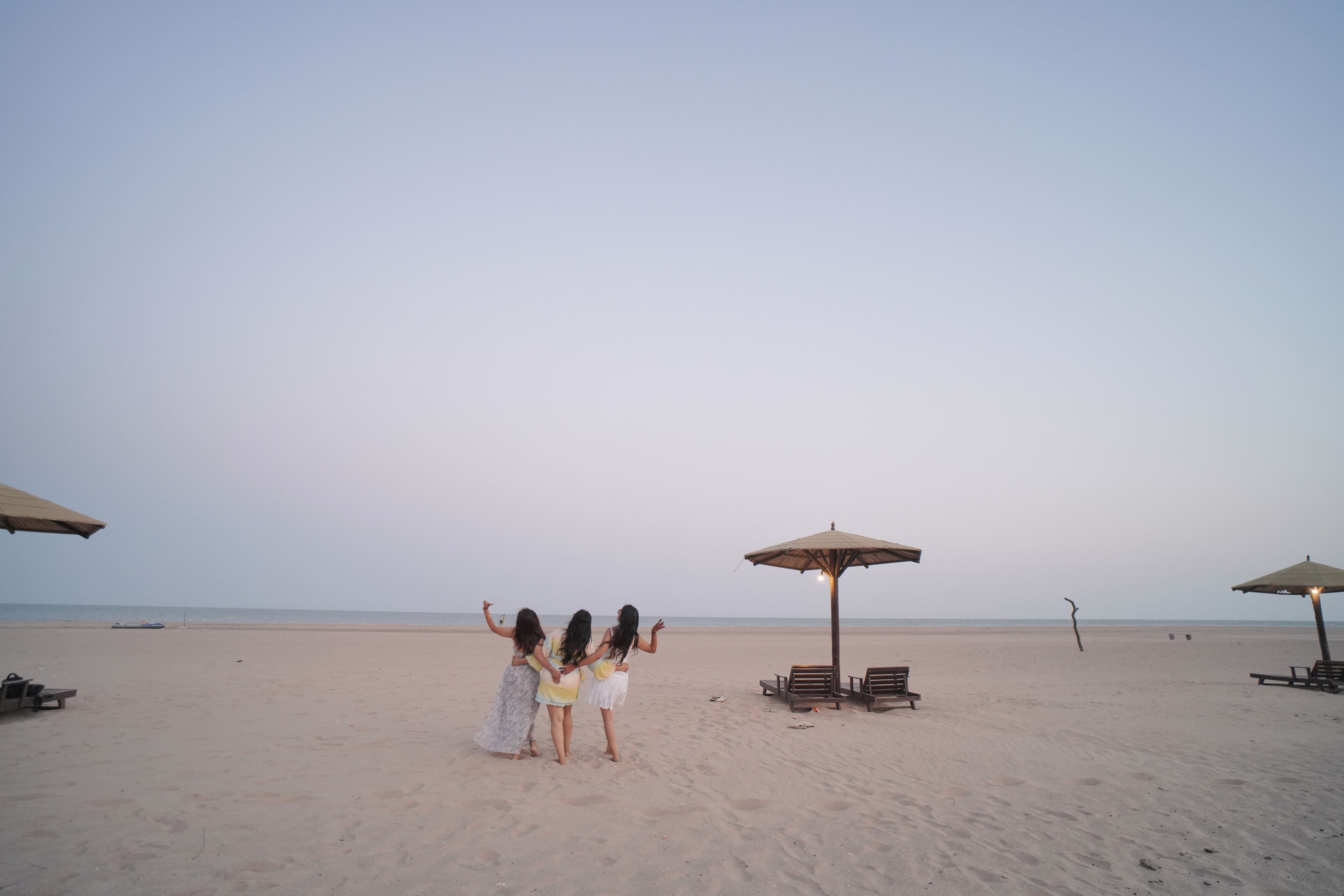 A beach with three people standing and facing the ocean, surrounded by beach umbrellas and lounge chairs.