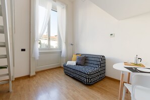 A room with a patterned sofa, a dining table with chairs, a window with curtains, and a thermostat on the wall.