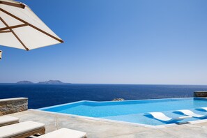 Céu azul claro, piscina com vista para o mar e uma parede de pedra.