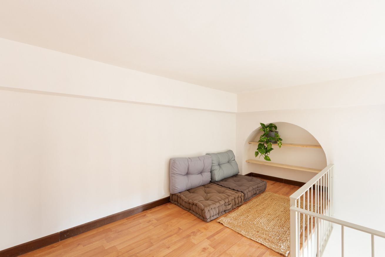 A corner room with a wooden floor, a sofa with cushions, a hanging plant, and a white railing.