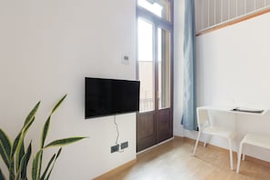 A room with a flat-screen TV mounted on the wall, a potted plant, a white chair, and a small table with a wine glass.