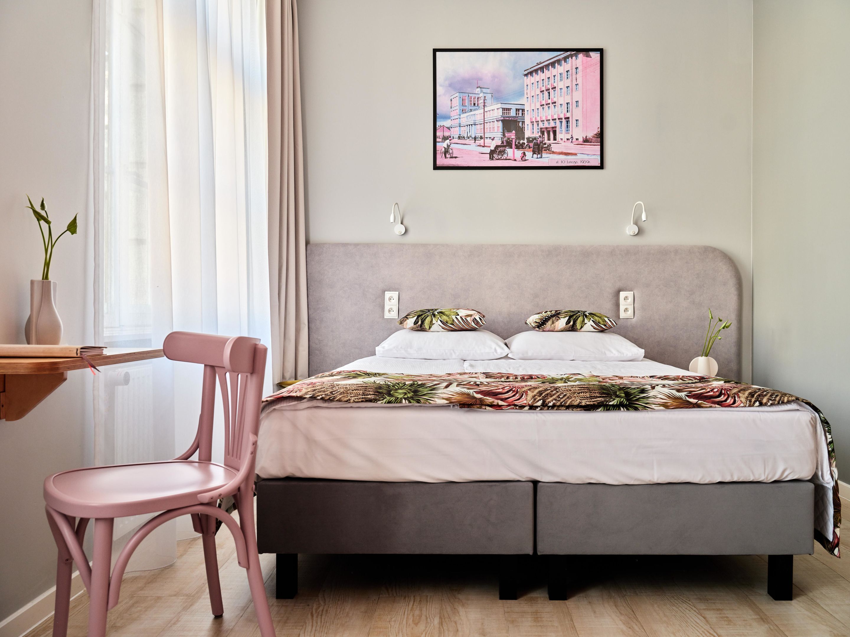 Un dormitorio con cama, una silla, mesita de noche, ventana con cortinas e imagen colgada en la pared.