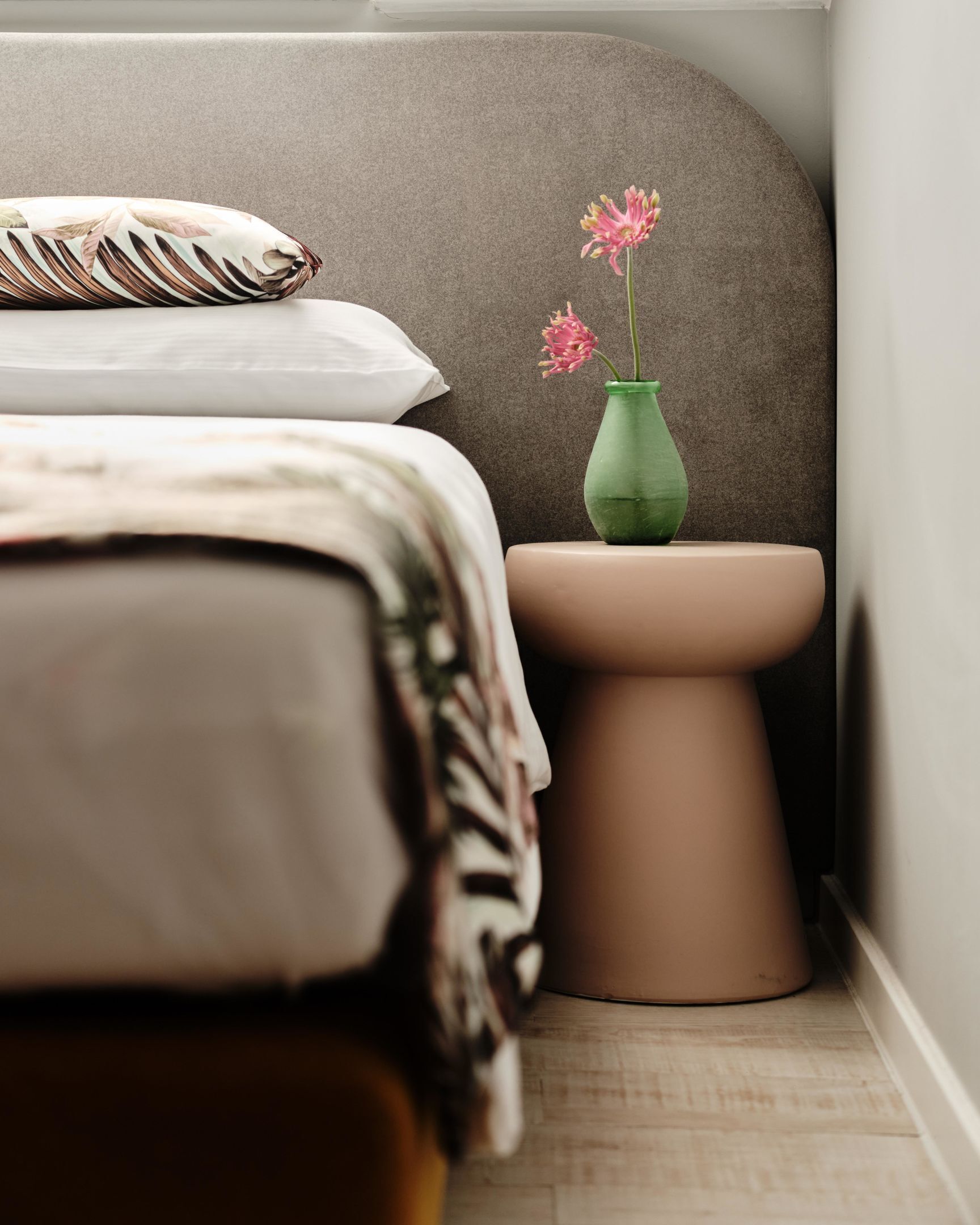 Un dormitorio con una cama, almohadas, una mesita de noche y un florero con flores.