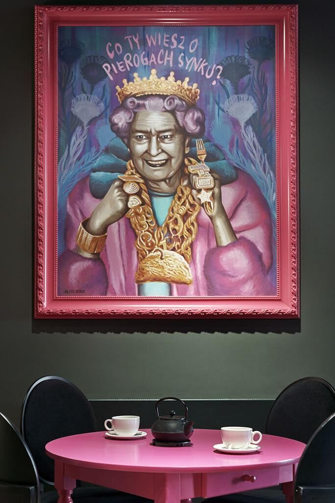 Un retrato enmarco de una persona vestida con atuendo real, con corona y cetro, sobre un fondo floral. Debajo del retrato, hay una mesa rosa con dos tazas blancas y una tetera.