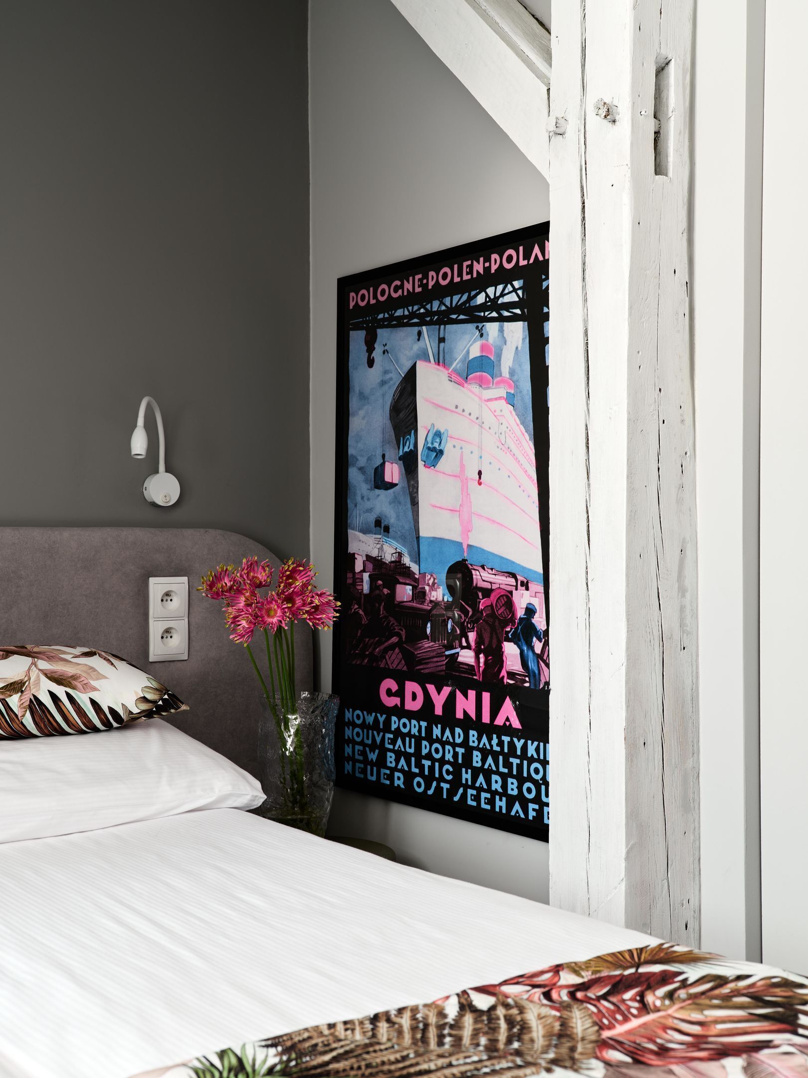 A bedroom with a bed, a nightstand, a wall-mounted lamp, a poster, and a vase with flowers.