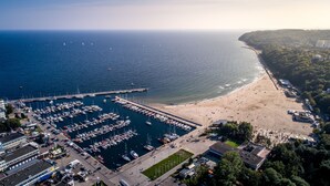 An aerial view of a marina with numerous boats docked, a sandy beach, and a forested hillside in the background. - Hola Gdynia (Gdynia)