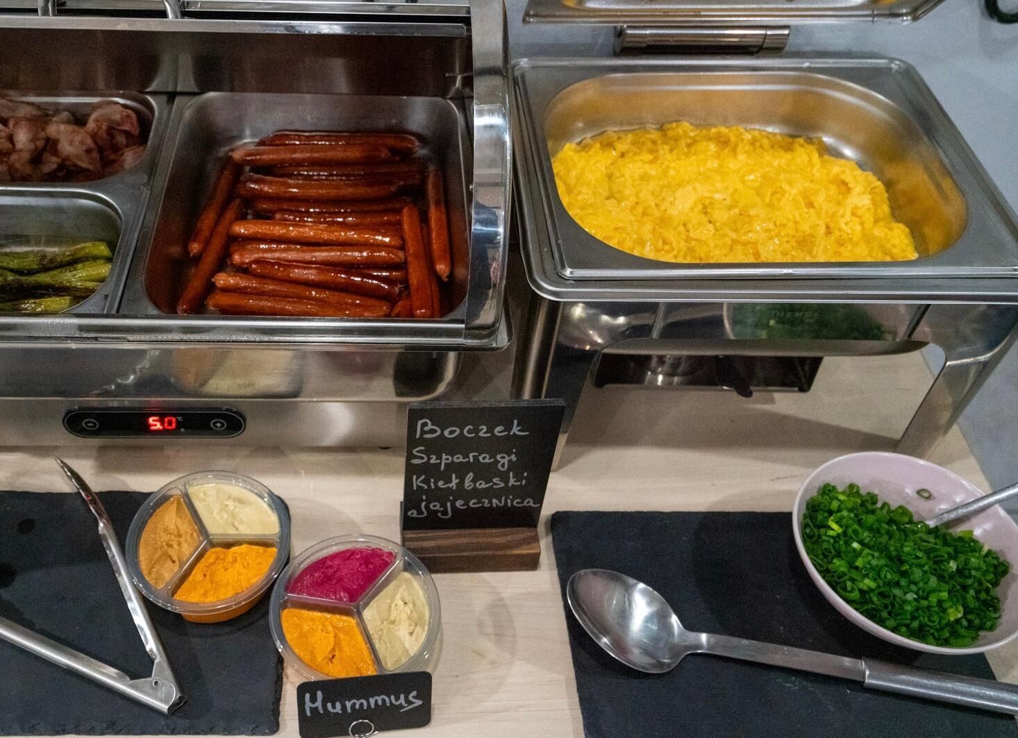 A buffet with various food options including sausages, scrambled eggs, hummus, and a bowl of green onions.