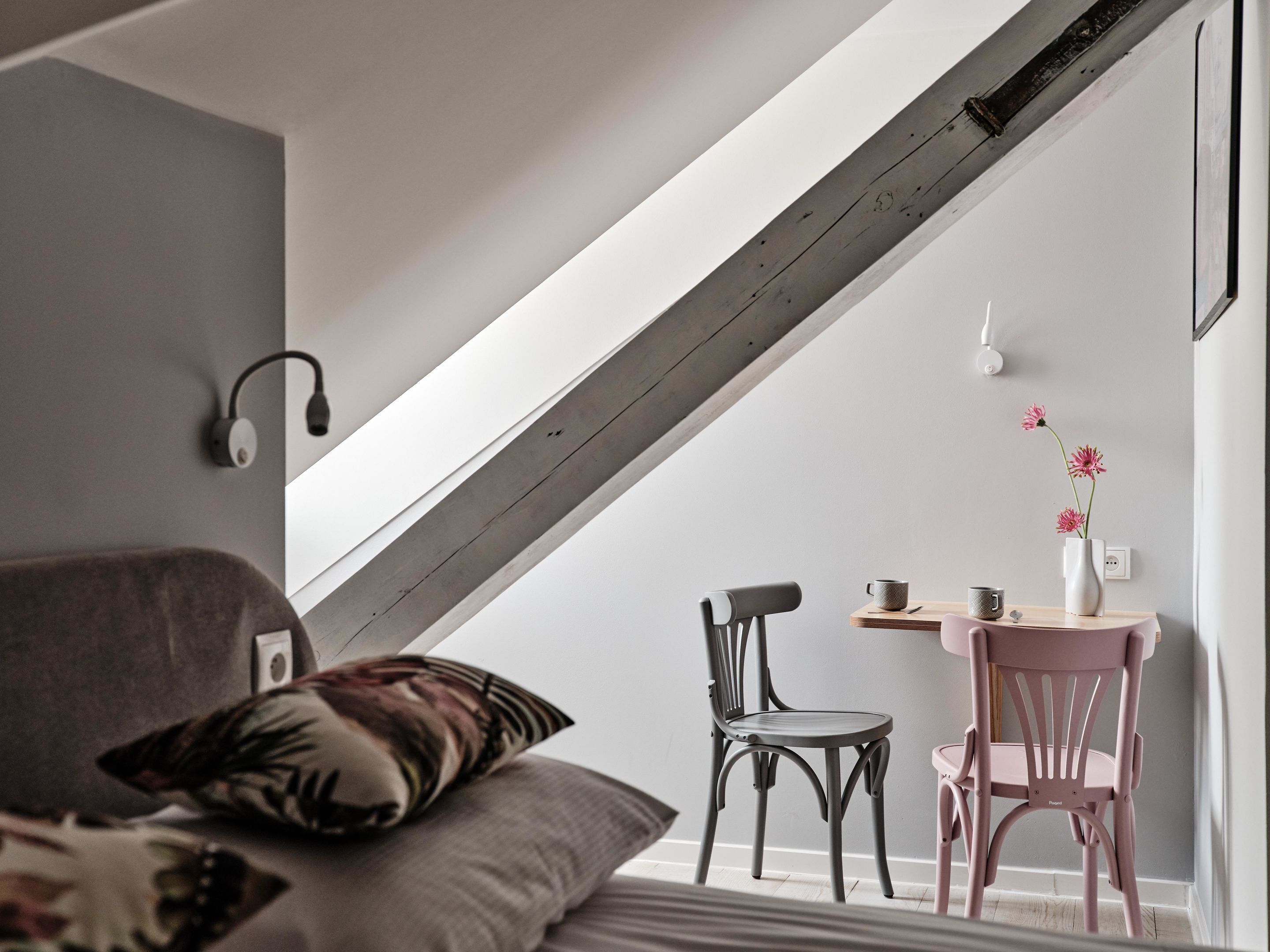 Un dormitorio estilo loft moderno con techo inclinado, cama con cojín estampado, escritorio con silla y un florero con flores.
