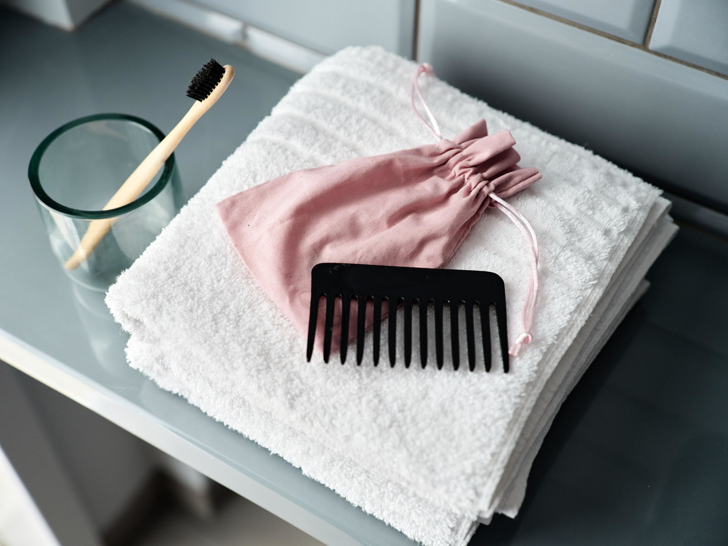 A bathroom with a white towel, a pink cloth, a black comb, and a wooden-handled toothbrush on a glass-topped table.