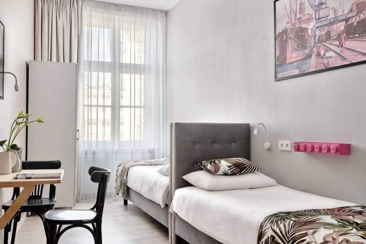A bedroom with a bed, a desk, a chair, a window with curtains, and a wall-mounted light.