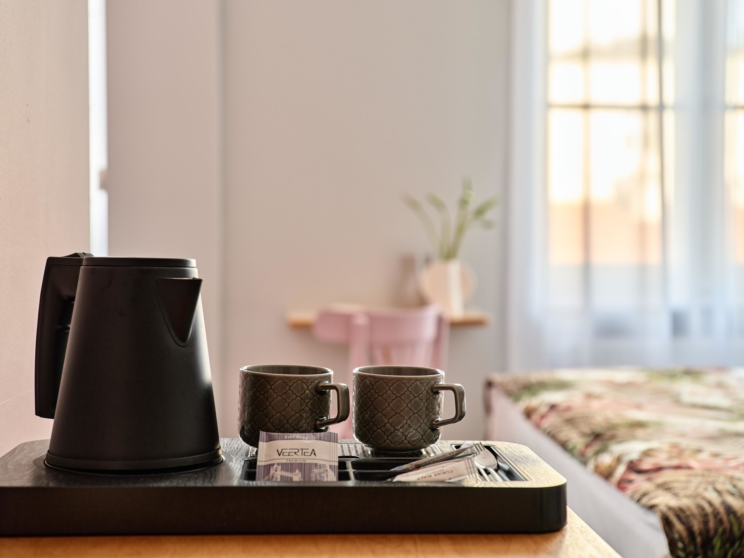 A tray with a black coffee maker, two patterned mugs, and a tea packet labeled "VERTEA" on a wooden table in a room with a bed and a window.