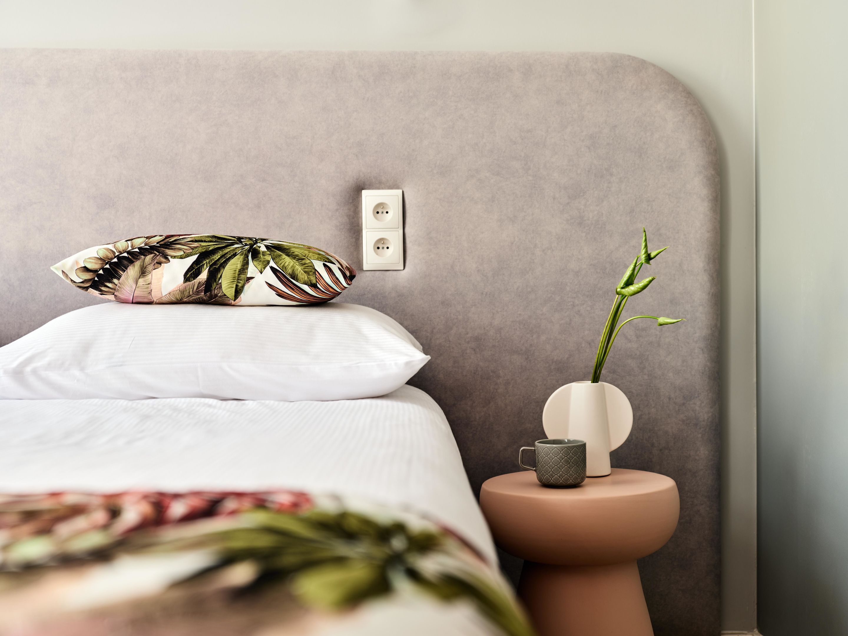 Un dormitorio con una cama, una mesita de noche, un florero con una planta y un enchufe en la pared.