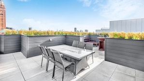 En takterrass med ett glasbord och flätade stolar, omgiven av grå väggar och utsikt över stadens silhuett.