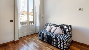 Una habitación con un sofá estampado, piso de madera, una puerta blanca con vistas y una lámpara fijada a la pared.