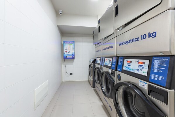 Un espace buanderie équipé de plusieurs machines à laver, chacune portant l’étiquette « Sèche-linge 10 kg », ainsi qu’un panneau de commande et un panneau indiquant « Point de lavage ».