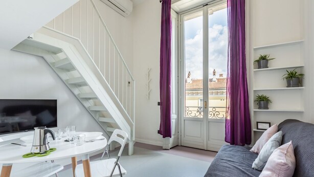 Una habitación moderna con una escalera blanca, un área de comedor con una mesa y sillas, un sofá con cojines y un balcón con cortinas moradas.