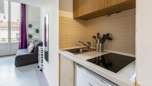 Cocina compacta con lavabo, estufa y armarios, junto a un balcón con cama y vistas a edificios.