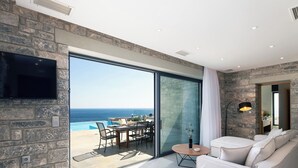 A modern living room with a stone wall, a flat-screen TV, a sofa, a small table, and a large sliding glass door leading to an outdoor patio with a pool and chairs.