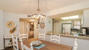 A dining area with a wooden table, white chairs, a chandelier, and a kitchen with modern appliances.