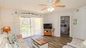 A living room with a ceiling fan, a TV on a wooden stand, a sofa, a coffee table, and a view of a bedroom through a sliding door.