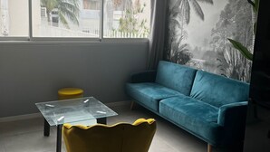 Una sala de estar con un sofá azul, un sillón amarillo, una mesa de centro de vidrio y un ventanal con vistas a palmeras y edificios.