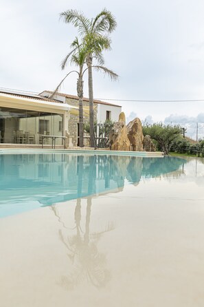 A swimming pool with clear water, surrounded by a house with a tiled roof, palm trees, and large rocks. - La Corte Del Sole (Castellammare del Golfo)