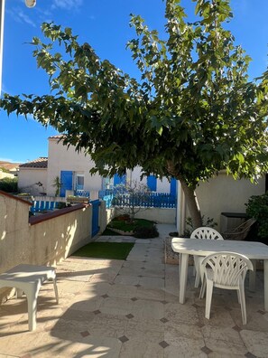 Uma área de varanda externa com piso de azulejos, cadeiras de plástico branco e uma pequena mesa.