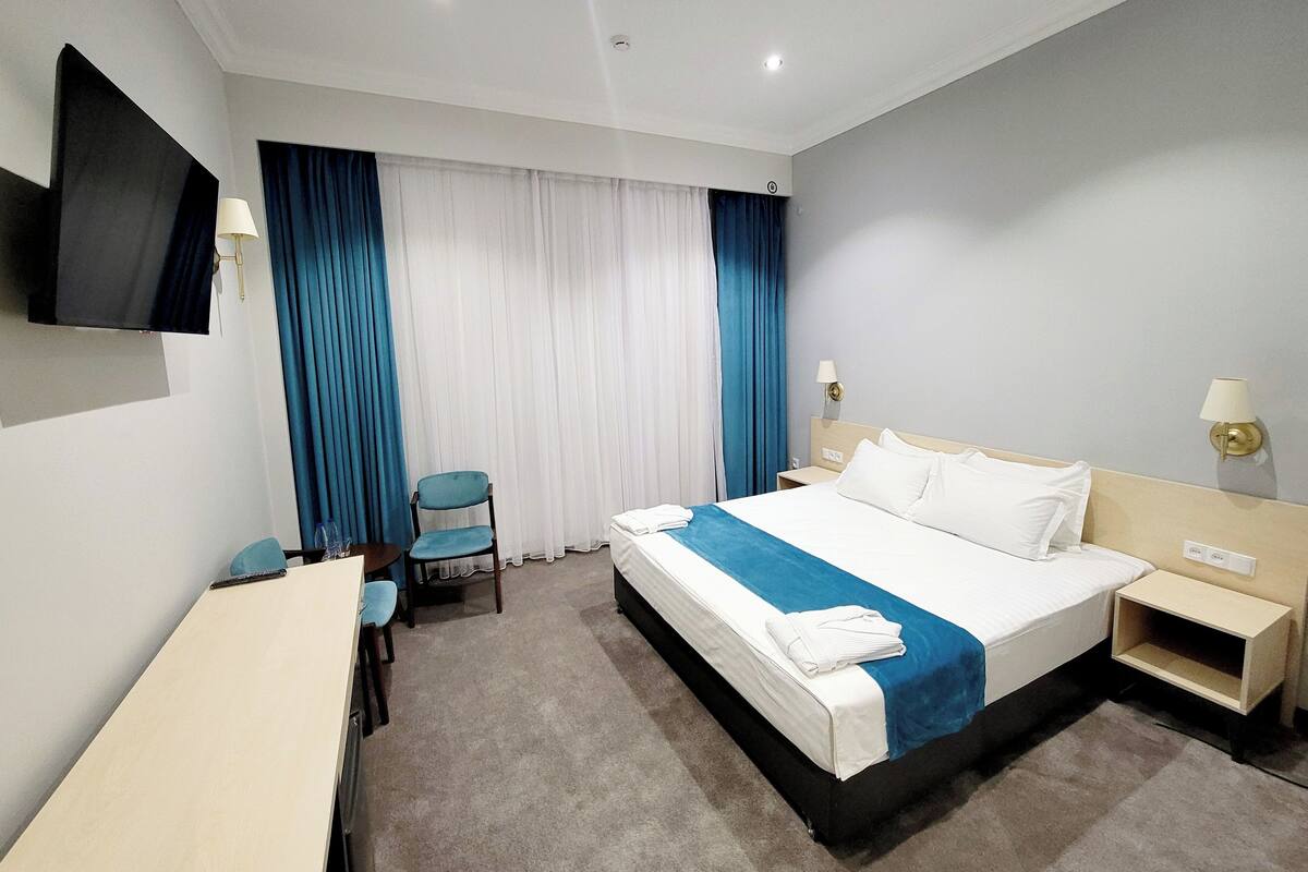 A hotel room with a bed, a desk, a chair, a TV, and a window with curtains.
