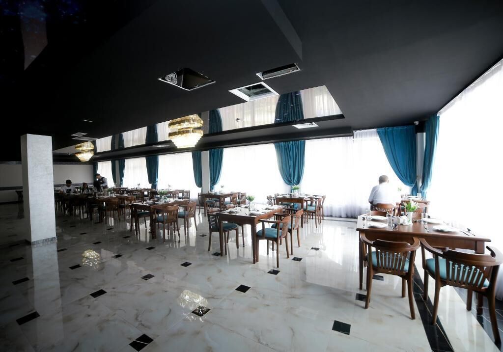 Un vaste espace dédié à la salle à manger, avec de nombreuses tables et chaises, de grandes fenêtres ornées de rideaux bleus et un lustre moderne.