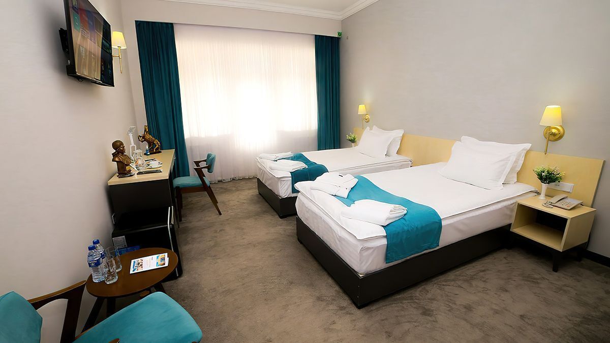 A hotel room with two beds, a desk with a chair, a small table with a chair, and a nightstand with a lamp.
