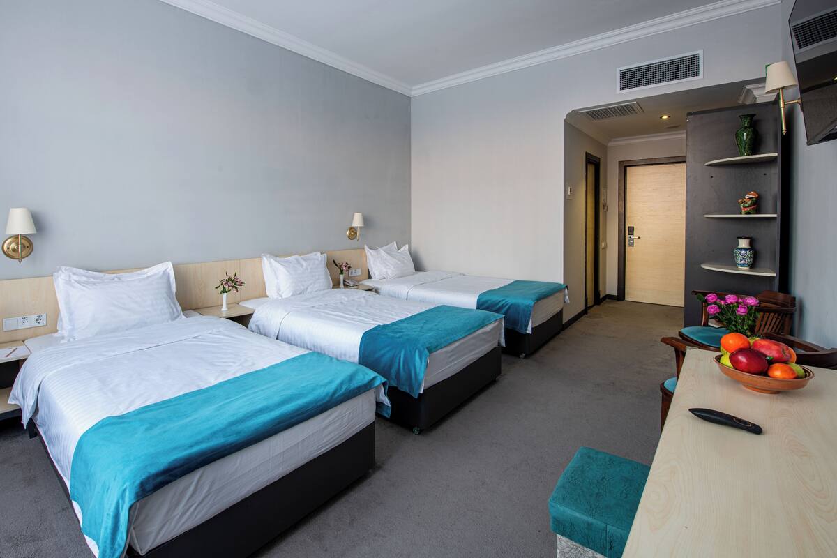 A hotel room with two beds, a desk with a bowl of fruit, a lamp, and a vase on a shelf.