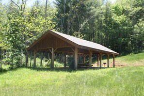 Un pavillon en bois doté d’un toit à pente, entouré d’une zone herbeuse et d’une forêt dense.
