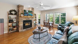 Un salon doté d’une cheminée en pierre, d’un téléviseur, d’un ventilateur de plafond, d’un coin repas avec des chaises et d’une grande fenêtre donnant sur les arbres.
