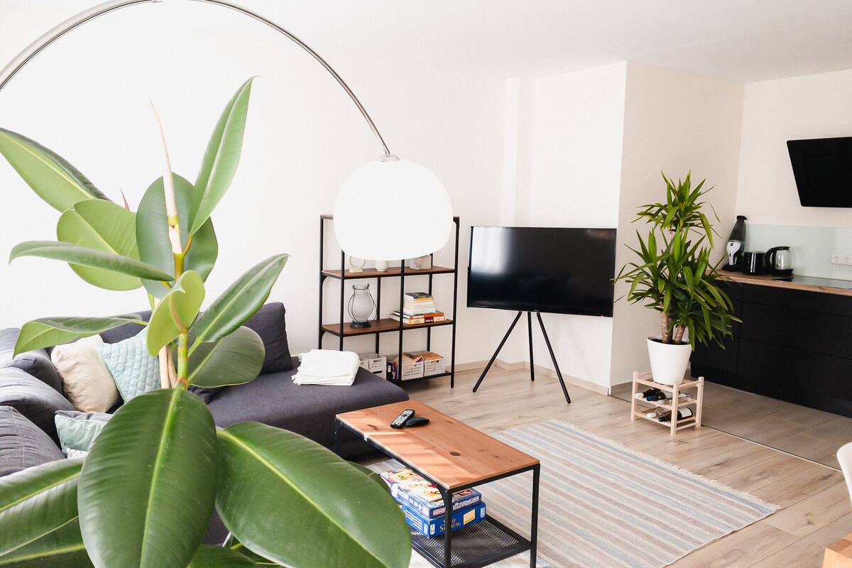Una sala de estar moderna con sofá, mesa de centro, televisor, estantería y plantas en macetas.