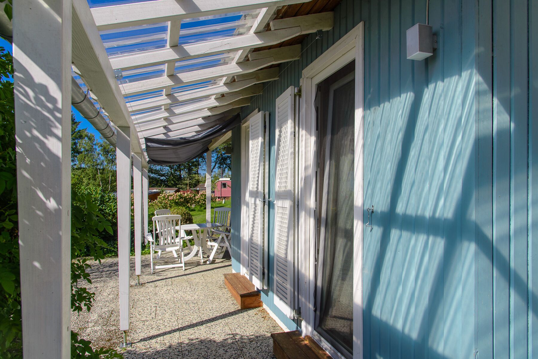 A covered porch with a table and chairs, a door, and a view of a garden and neighboring buildings.