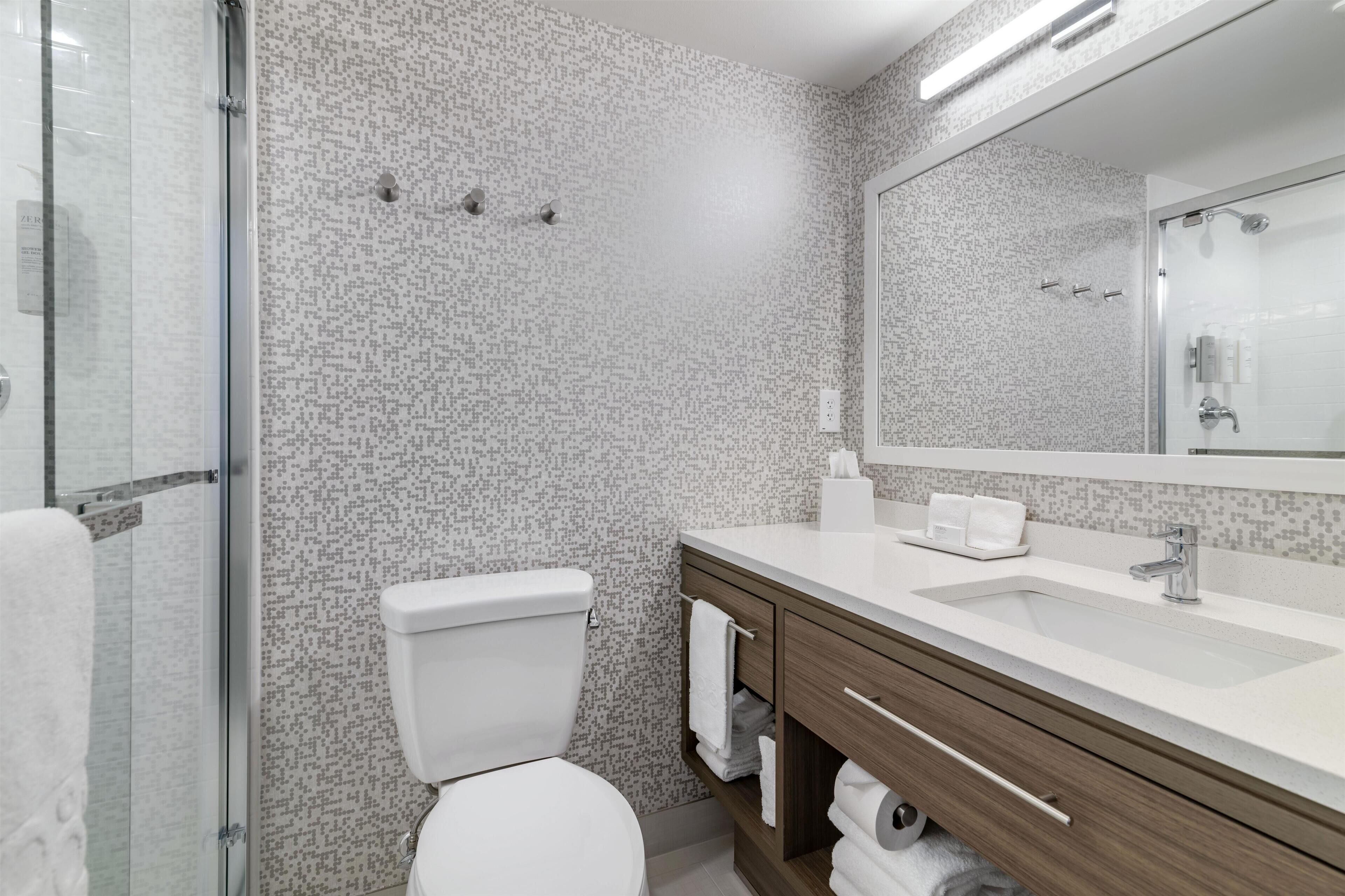 A modern bathroom with a patterned wall, a white toilet, a glass shower enclosure, and a wooden vanity with a sink and mirror.