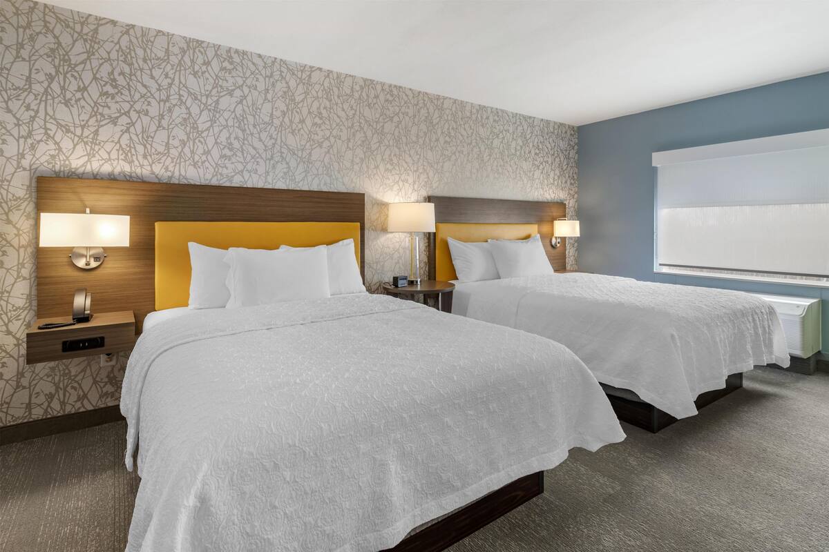 A hotel room with two beds, a patterned wallpaper, a blue accent wall, and a window with blinds.