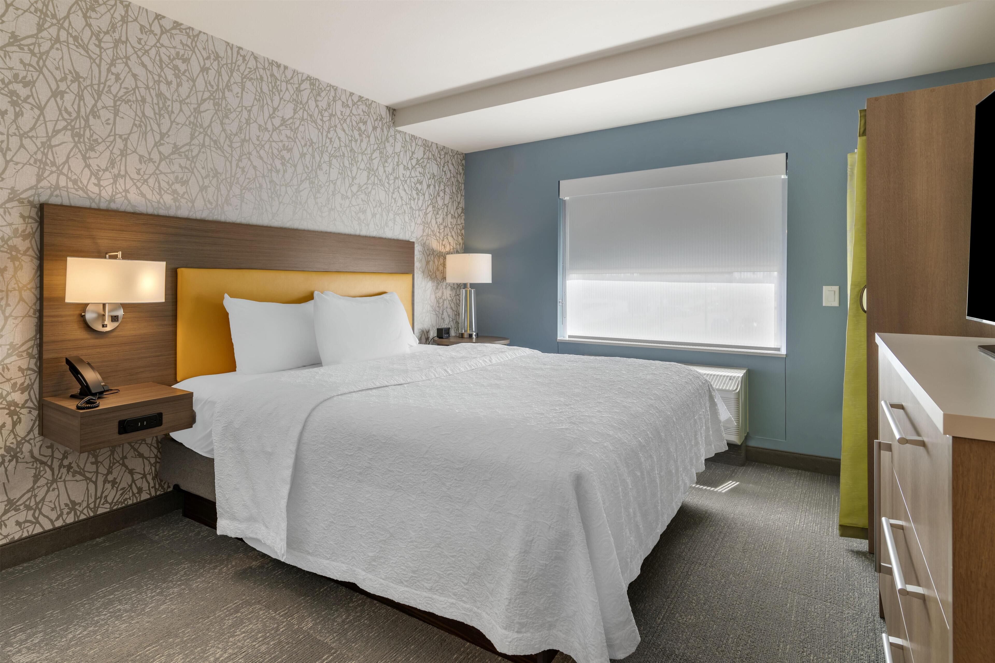 A hotel room with a bed, nightstands, a desk, a chair, a TV, and a window with blinds.